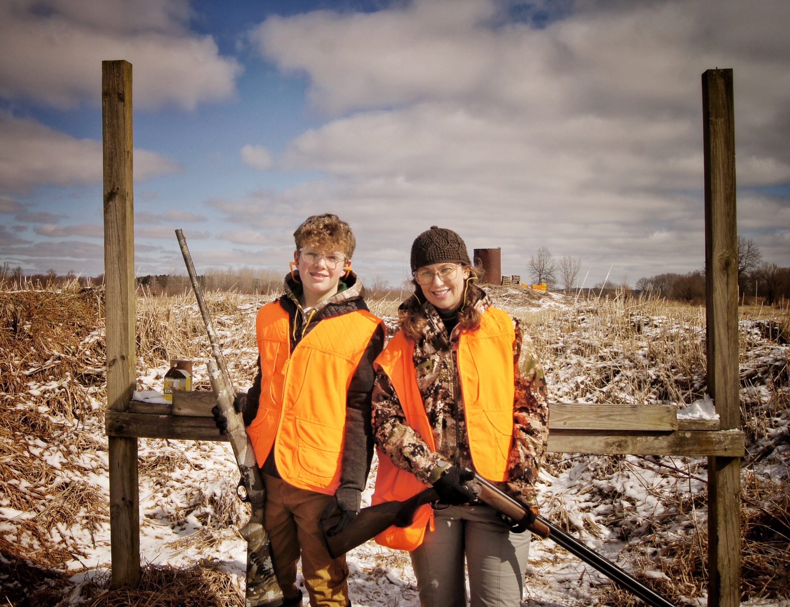 Two boys stand in an open field wearing hunting safety gear as they prepare for a pheasant hunt.