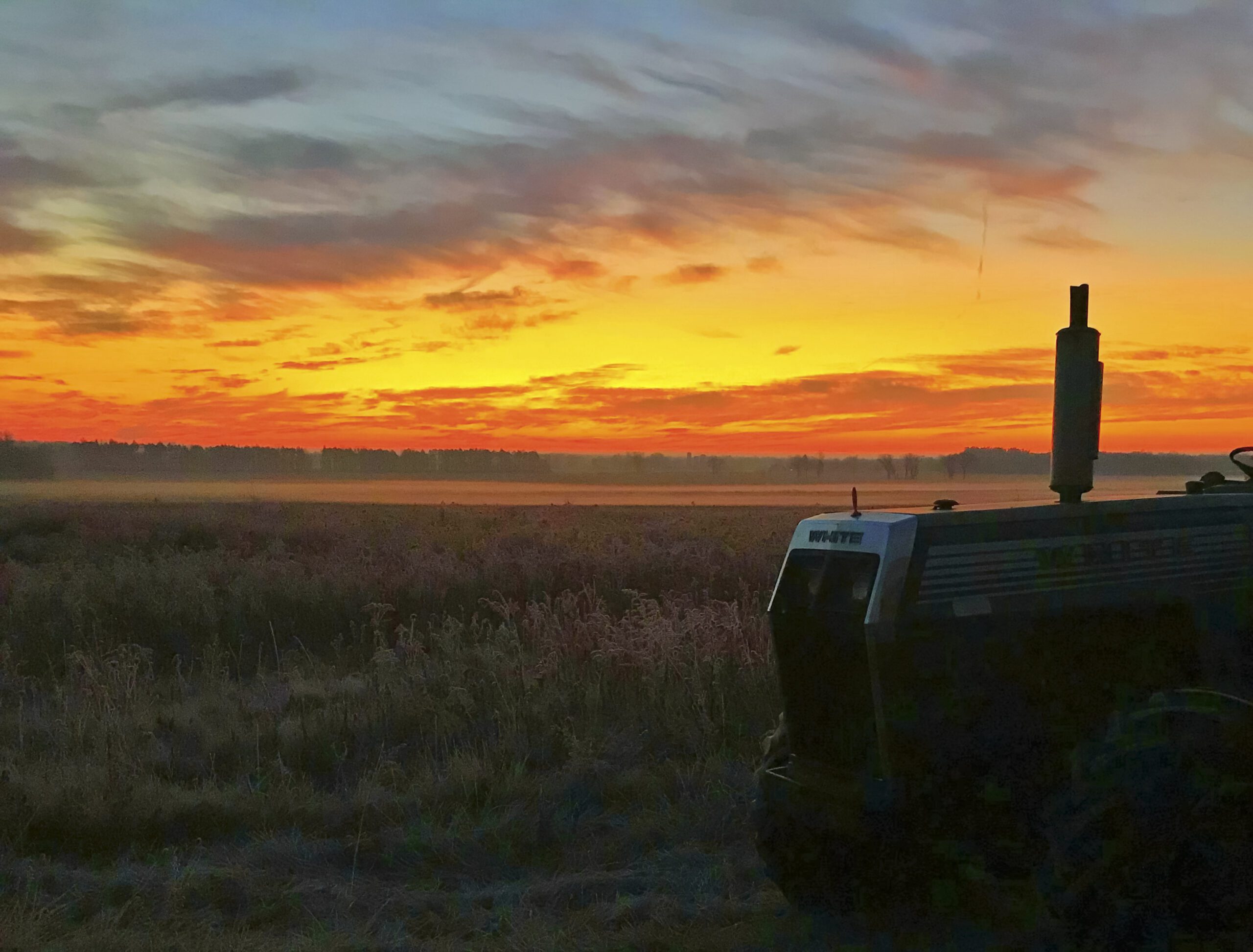 An idle tractor sits in a field as sunrise turns the skies vibrant colors of red, orange, and blue